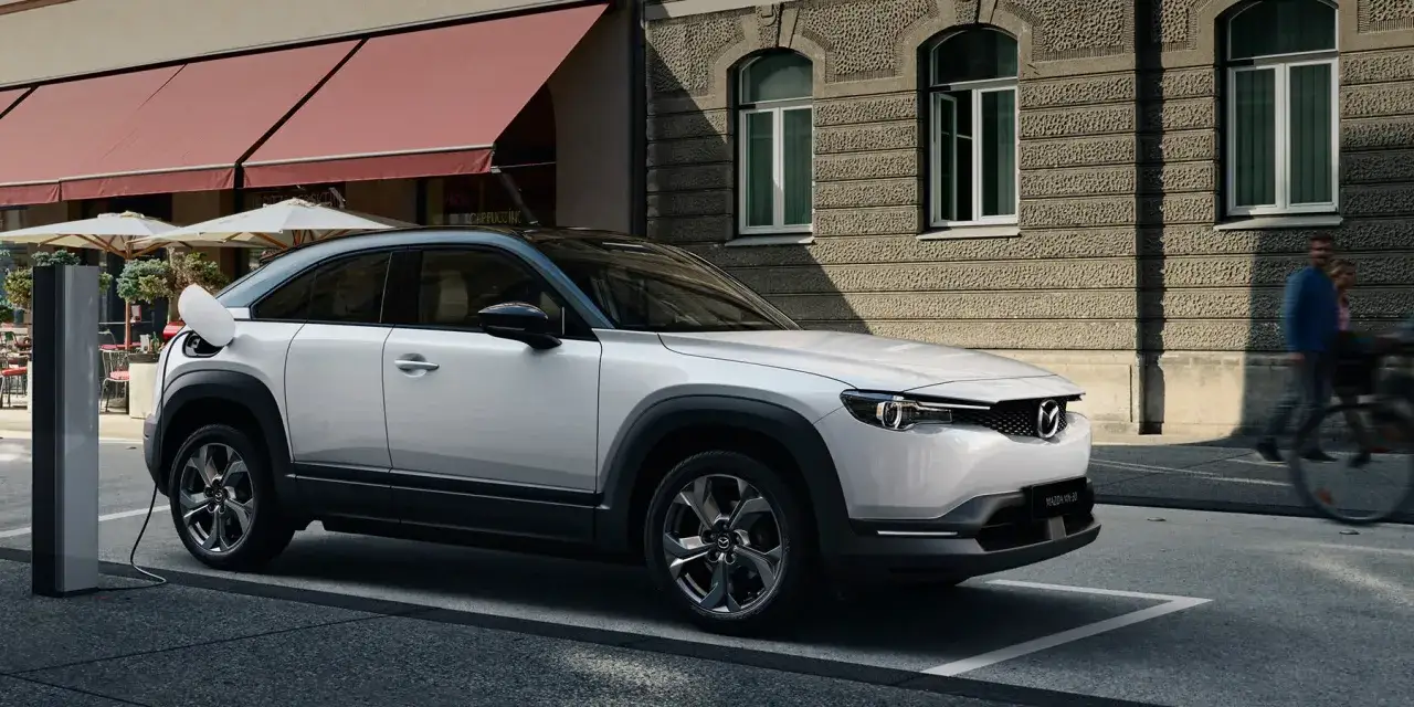 Biała Mazda MX-30 ładuje się przy miejskiej kawiarni. W tle widać ludzi spacerujących ulicą.