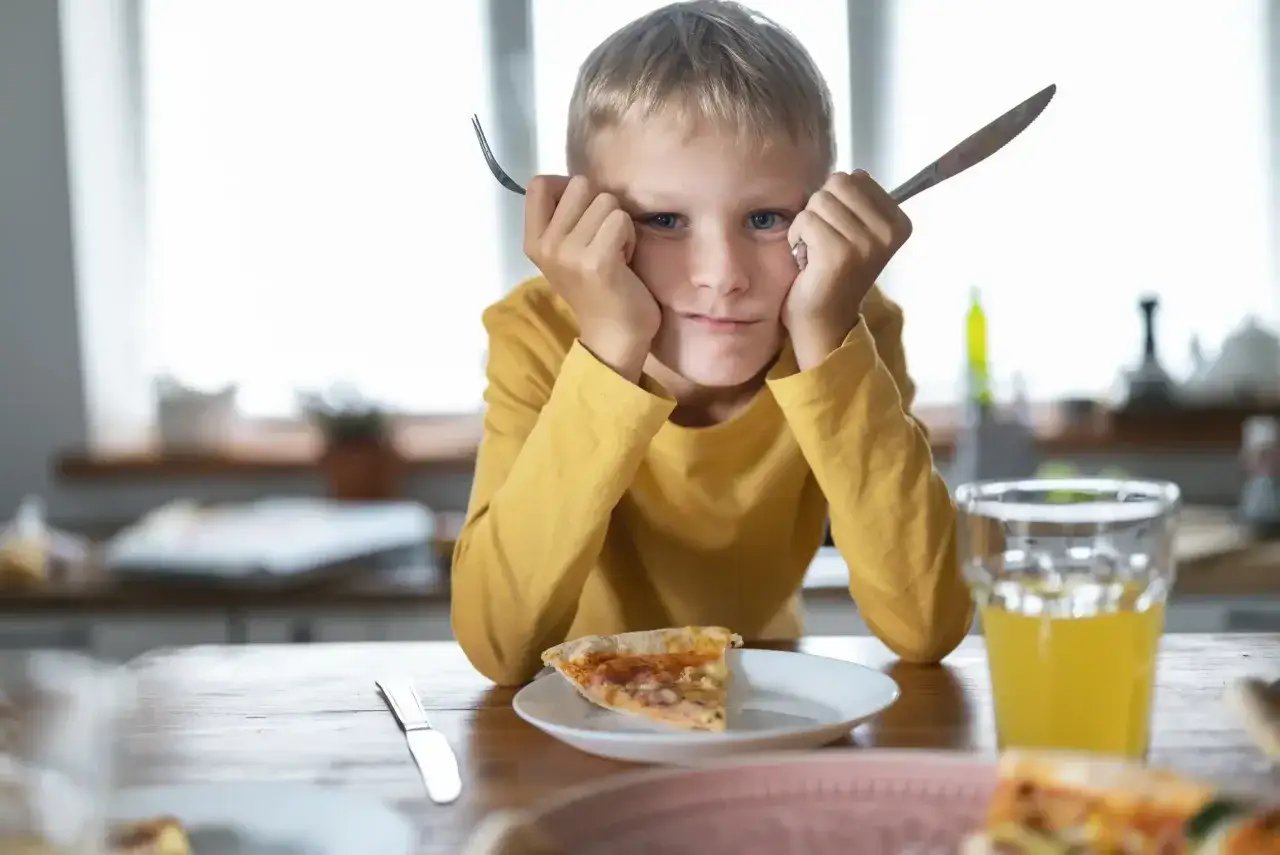 Chłopiec z widelcem i nożem w dłoniach, z miną wyrażającą znudzenie, siedzi przy stole z pizzą. Wygląda na typowego niejadka.