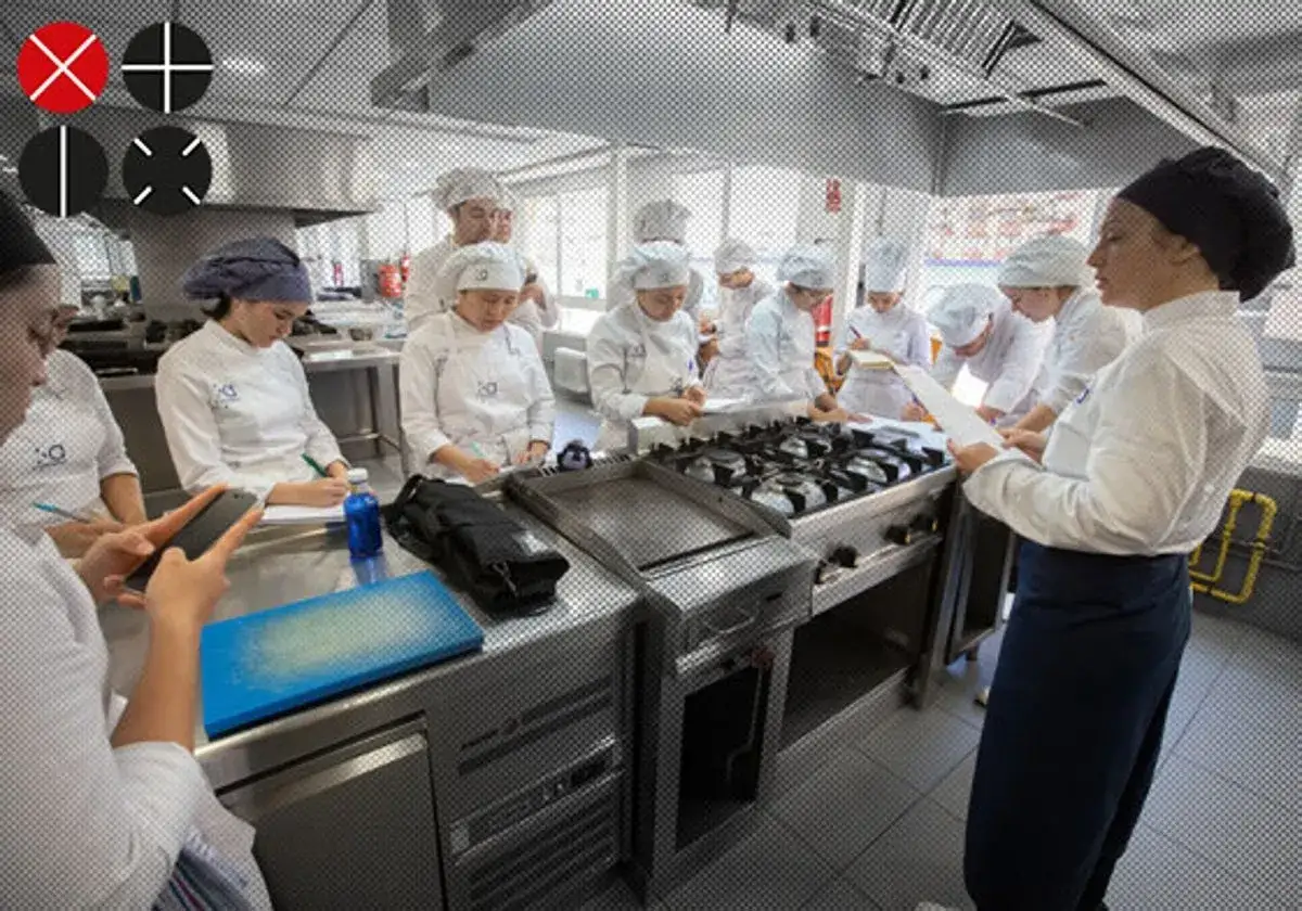 Estudiantes en un centro culinario mediterr&aacute;neo, aprendiendo y tomando notas en una cocina profesional.