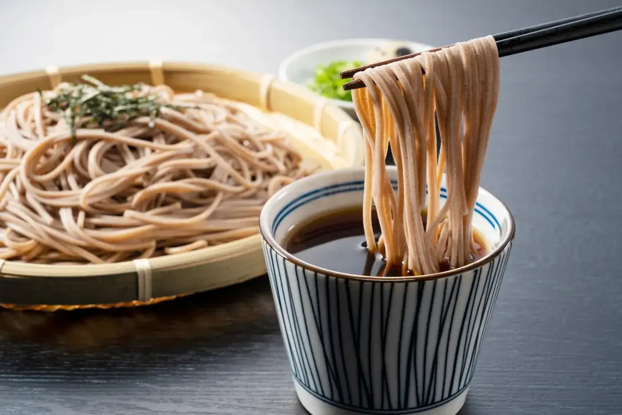 Makaron soba kontra makaron pełnoziarnisty. Pałeczkami podnoszone są nitki makaronu soba z miseczki z sosem.