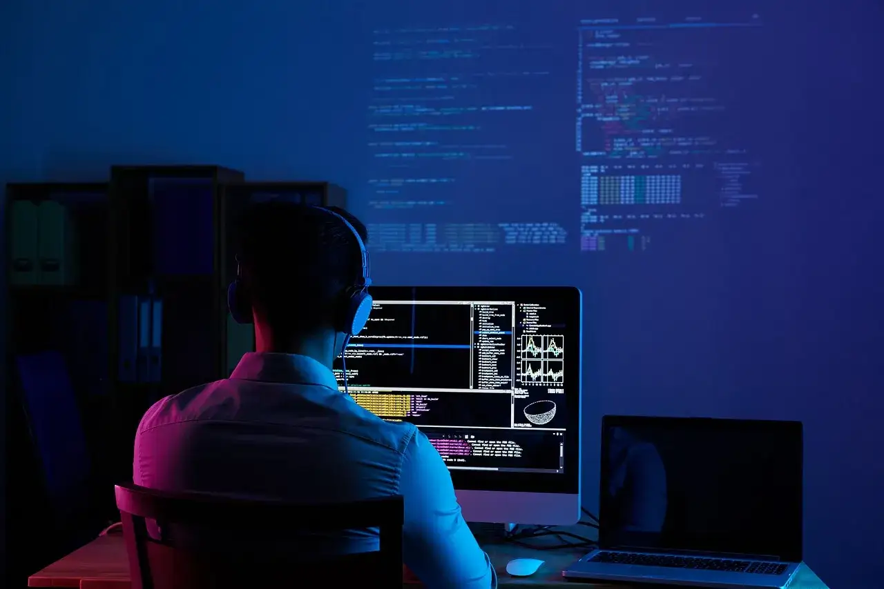 Programista w słuchawkach pochyla się nad monitorem, na którym widać linie kodu i wykresy. Na ścianie wyświetlają się kolejne kody PKD.