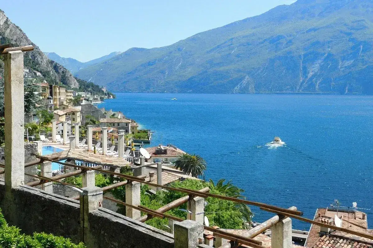 Vista sul lago con una barca, case colorate e una piscina. In primo piano, una struttura in legno ricorda una limonaia del castel.