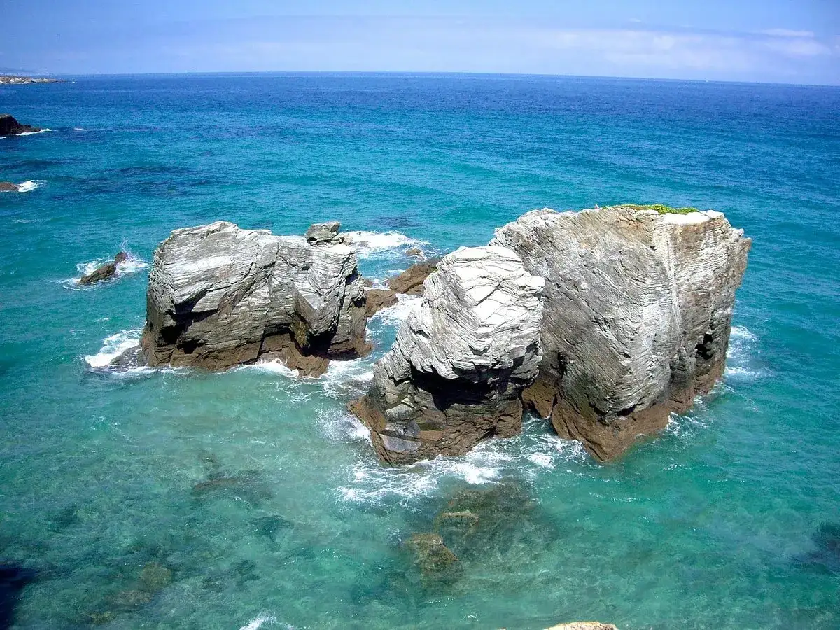 Impresionantes formaciones rocosas emergen del mar turquesa en la costa de Ribadeo, con olas rompiendo suavemente.