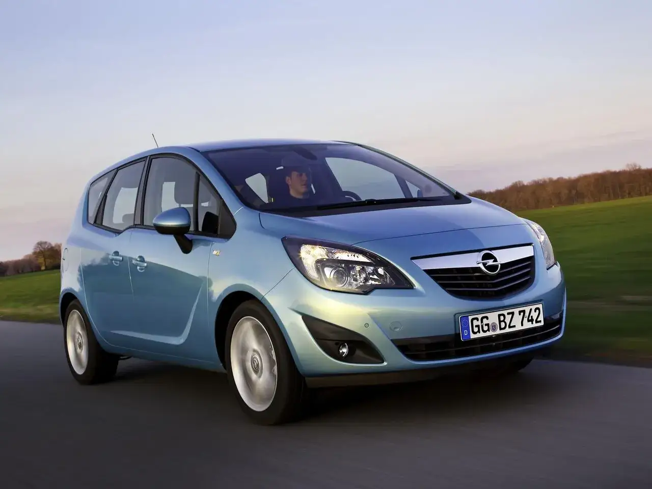 Błękitny Opel Meriva 1.6 benzyna w ruchu drogowym, z kierowcą za kierownicą, na tle krajobrazu.
