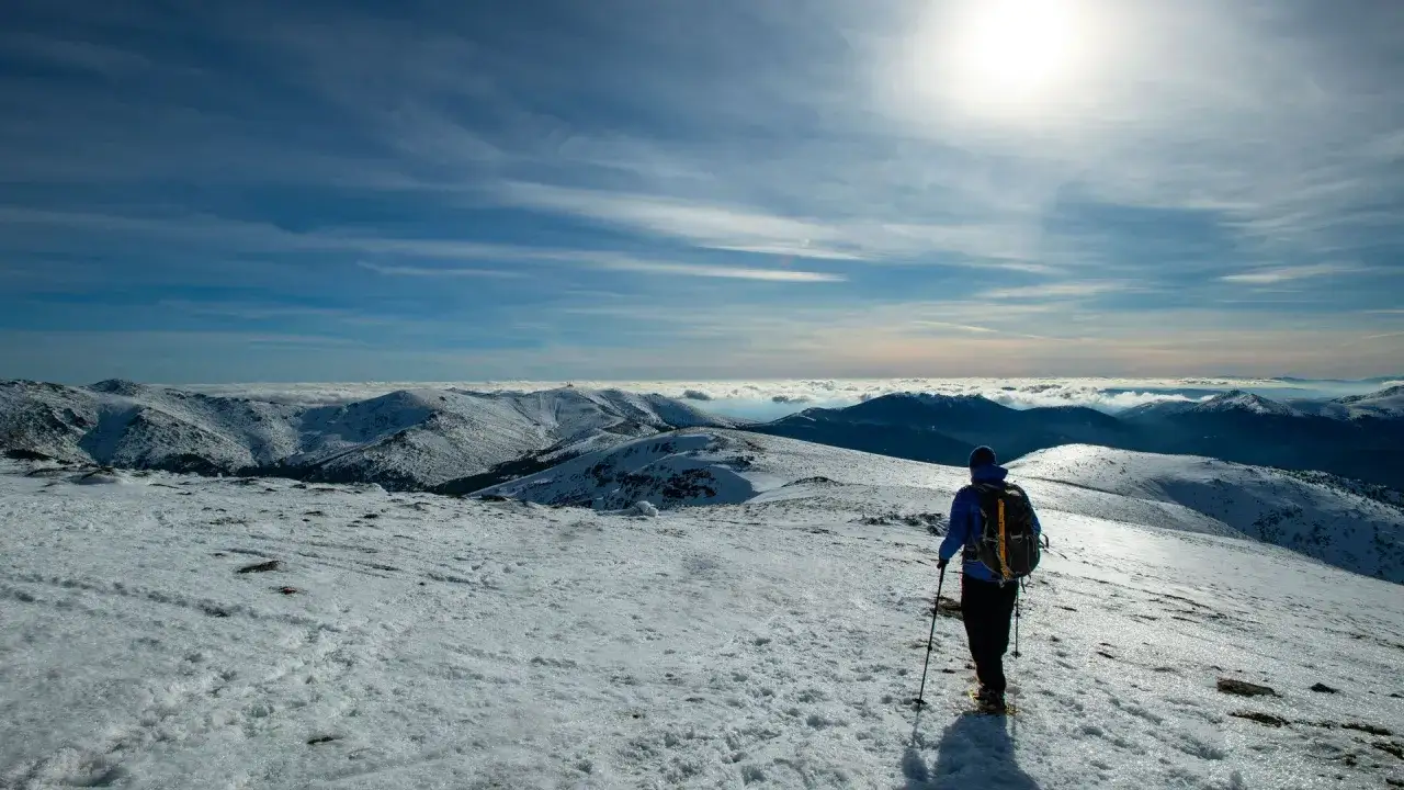 Senderismo en la **Peñalara** con **nieve**. Un excursionista con mochila y bastones contempla un paisaje montañoso cubierto de blanco bajo un cielo azul.