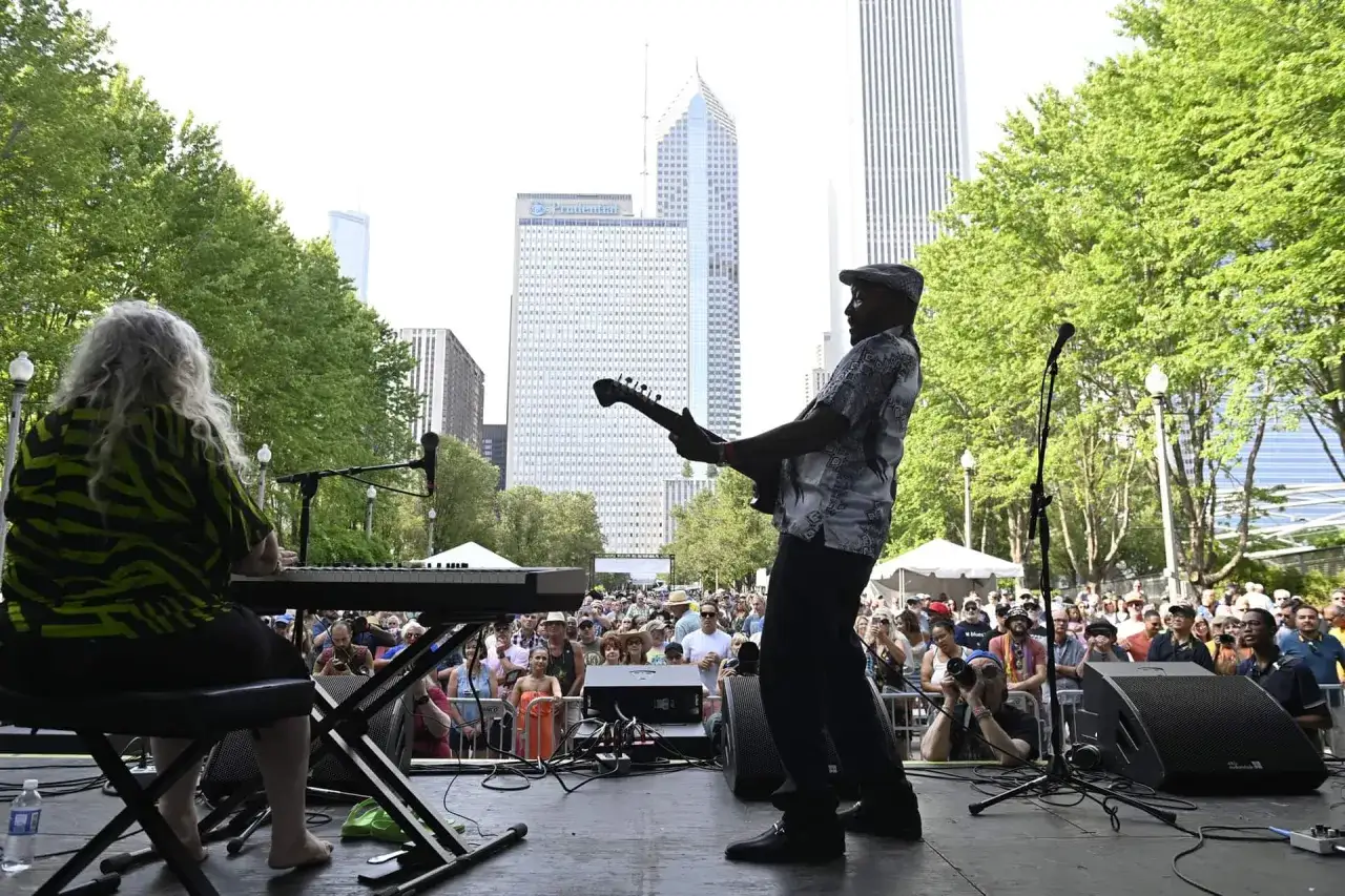 Un groupe joue du blues musique en plein air, avec des gratte-ciel en arrière-plan. Une pianiste aux cheveux longs et un guitariste en casquette sont sur scène.