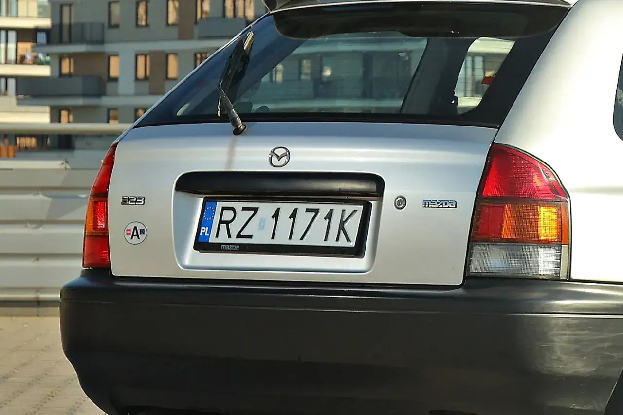 Tył srebrnej Mazdy 323 z tablicą rejestracyjną RZ 1171K. Widoczna naklejka z literą 