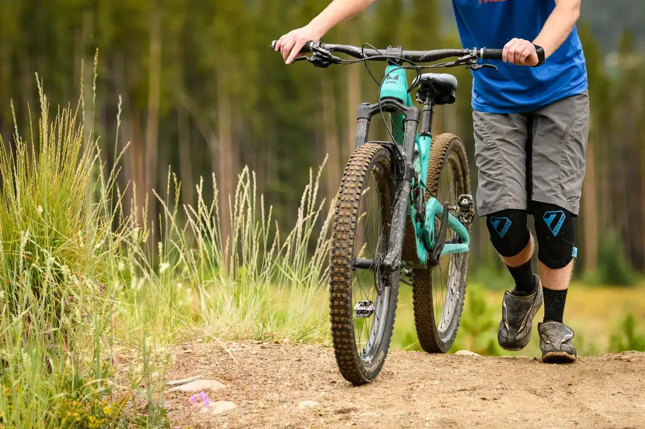 Person med mtb utrustning går längs en grusväg i skogen. Cykeln är turkos och lerig.