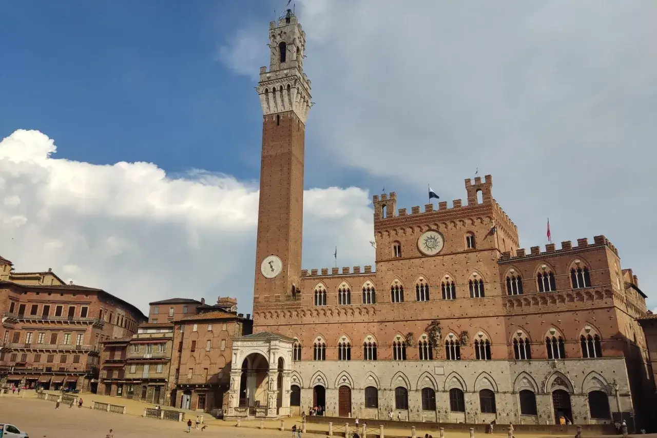 Wieża i Palazzo Pubblico w Sienie, z charakterystyczną ceglaną fasadą i kamiennymi detalami, pod błękitnym niebem z chmurami.