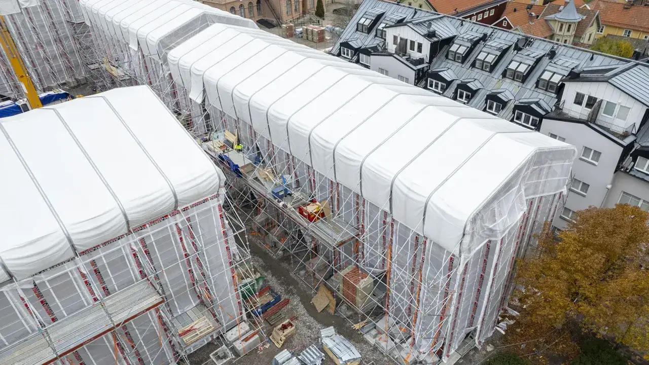 Byggarbetsplats i Sverige med vita presenningar över hus, ställningar och en gul kran. En pampuscher herr skulle uppskatta den ordning som råder.