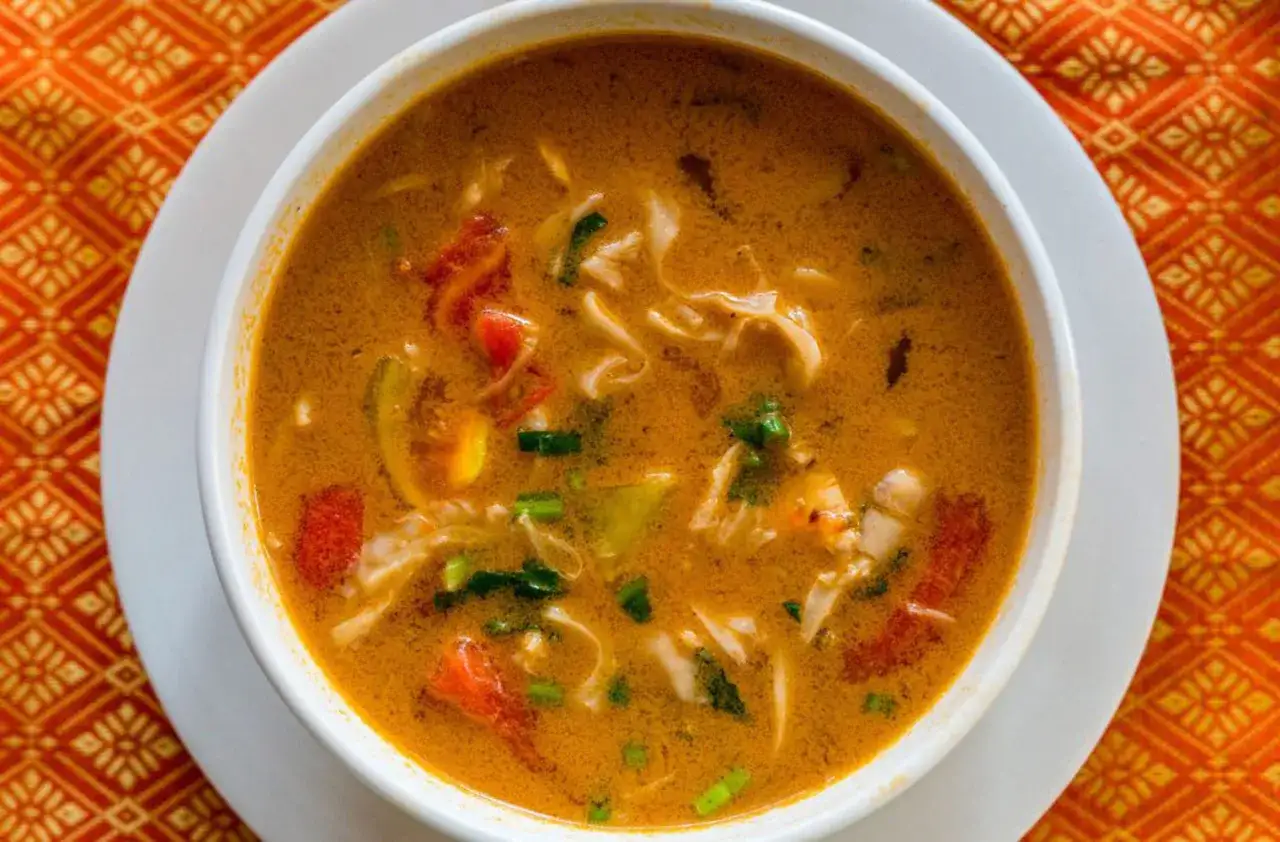 Aromaticzna zupa tom yum z kurczakiem, pełna kawałk&oacute;w kurczaka, pomidor&oacute;w i zi&oacute;ł, podana w białej misce na pomarańczowym, wzorzystym obrusie.