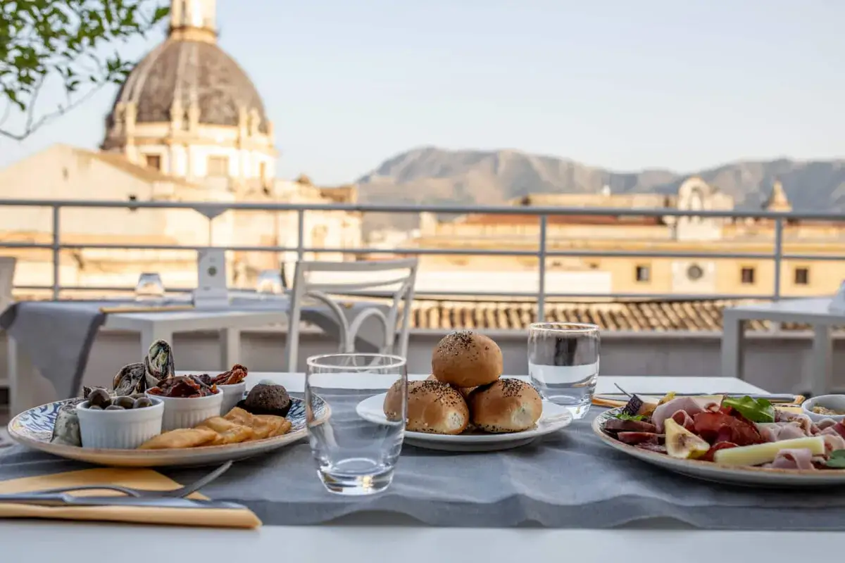 Un delizioso aperitivo a Palermo con taglieri di salumi, formaggi, olive e pane, con la cupola di una chiesa sullo sfondo.