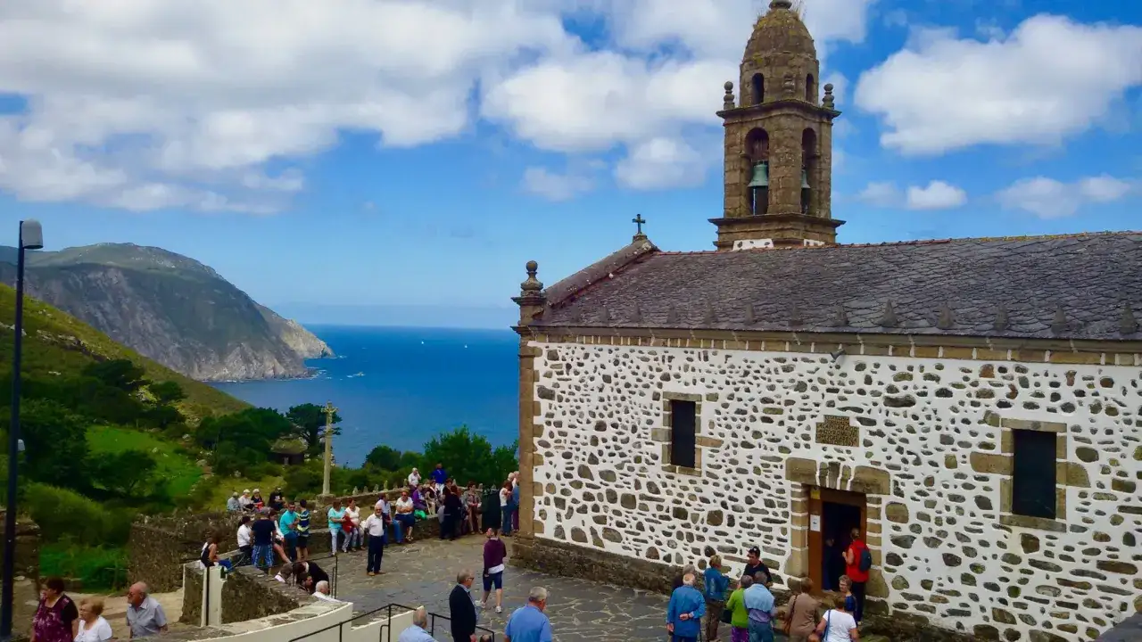 Iglesia blanca con torre campanario, rodeada de gente, con vistas a los acantilados de San Andrés de Teixido y el mar azul.