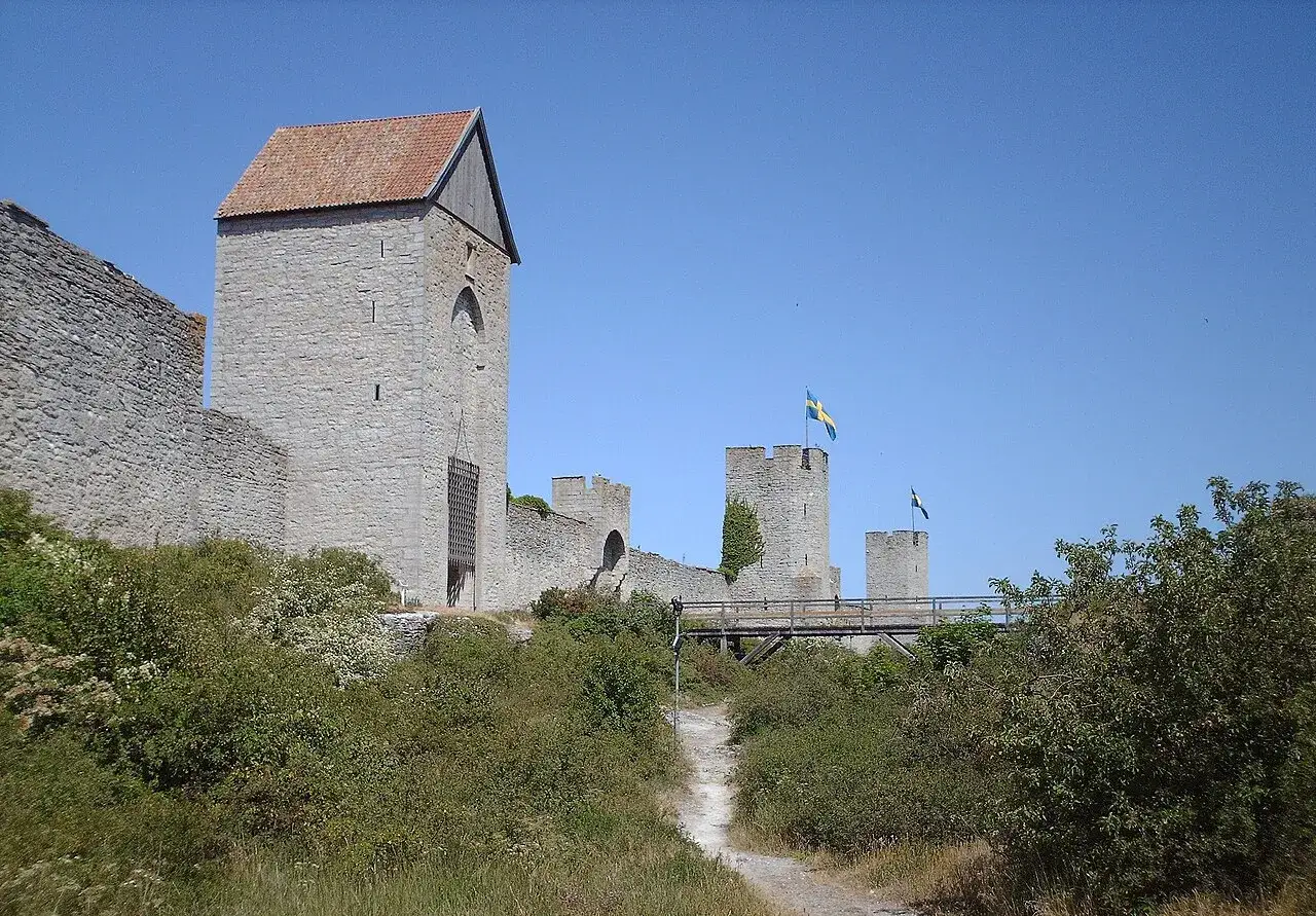 Imponerande ringmur med torn och svenska flaggor, en vacker vy att se i Visby. En stig leder upp mot en träbro.