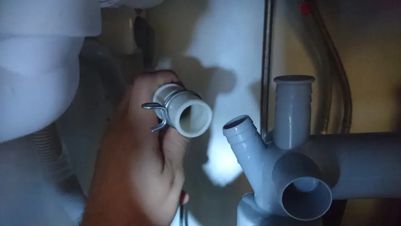 Kopplar slangen f&ouml;r vatten till diskmaskin. En hand h&aring;ller en vit slang&auml;nde med kl&auml;mma, redo att anslutas till ett gr&aring;tt avloppsr&ouml;r.