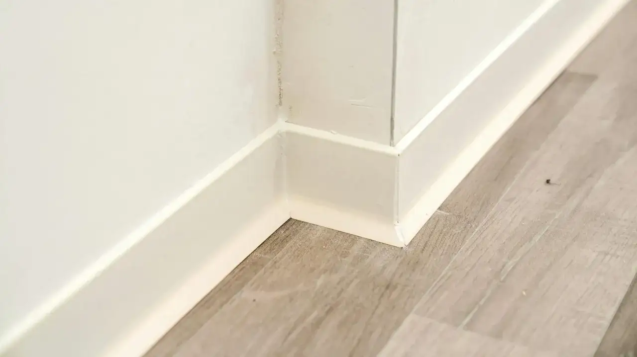 Angolo di una stanza con pavimento in laminato grigio e battiscopa bianco. La posa del battiscopa è curata, creando un raccordo pulito tra parete e pavimento.