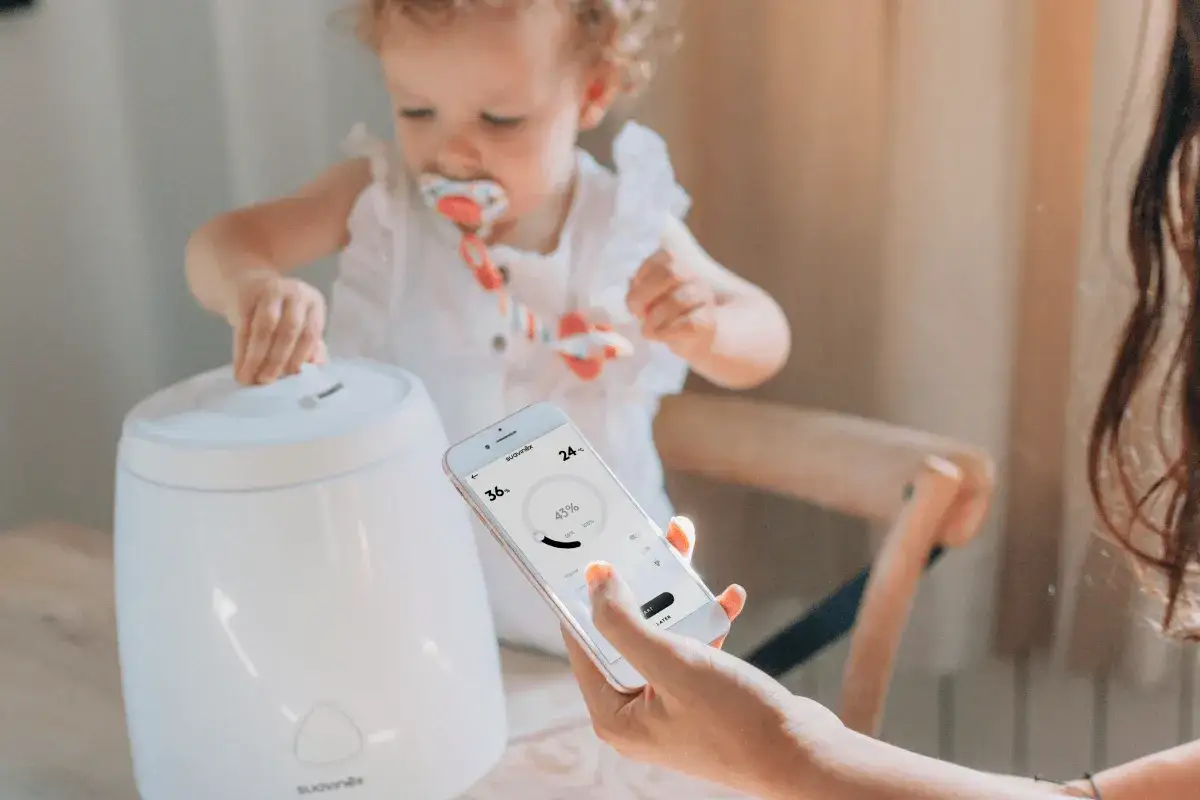 Un bebé juega cerca de un humidificador, mientras una mano controla el aparato desde un móvil. Este humidificador bebé para que sirve es ideal para crear un ambiente confortable.