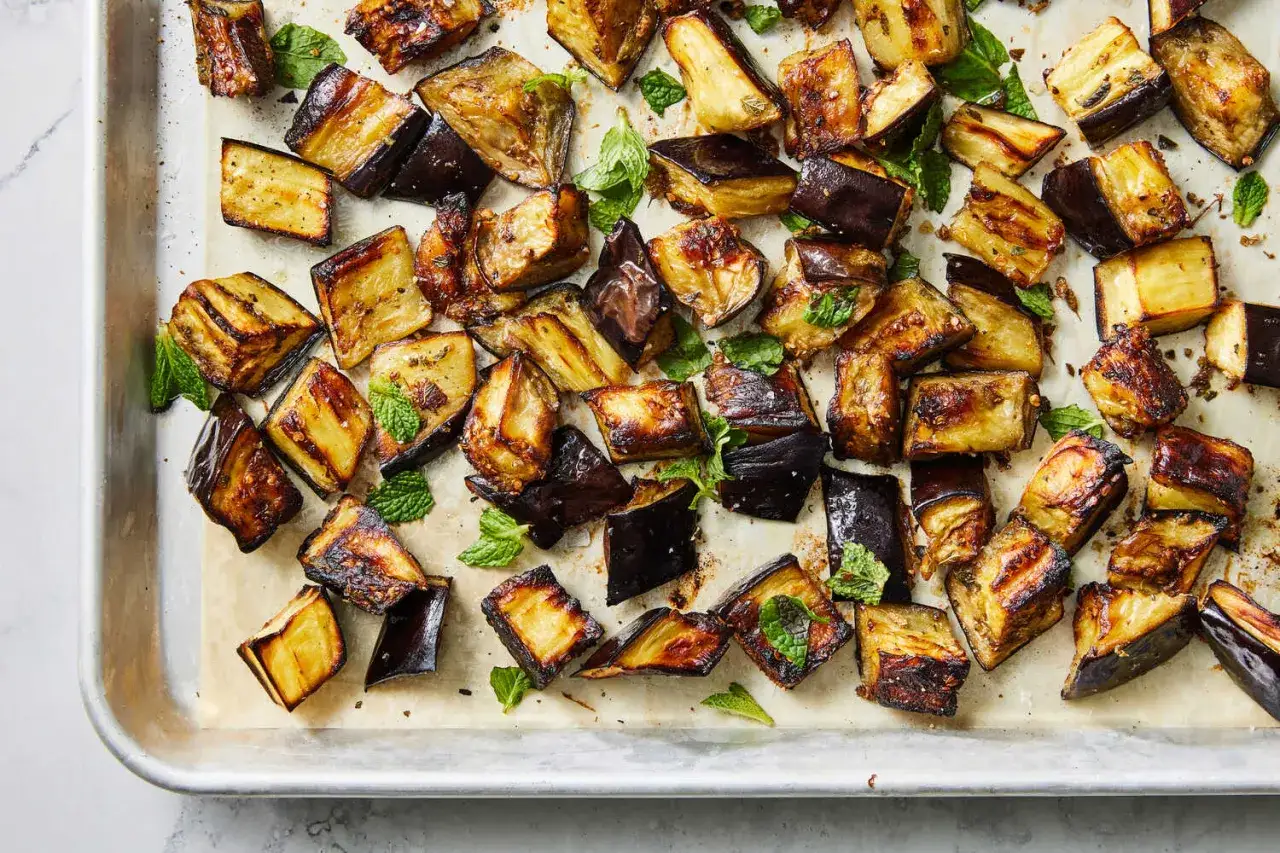 Ugnsrostad aubergine i bitar, gyllene och karamelliserade, strödda med färska myntablad på en plåt.