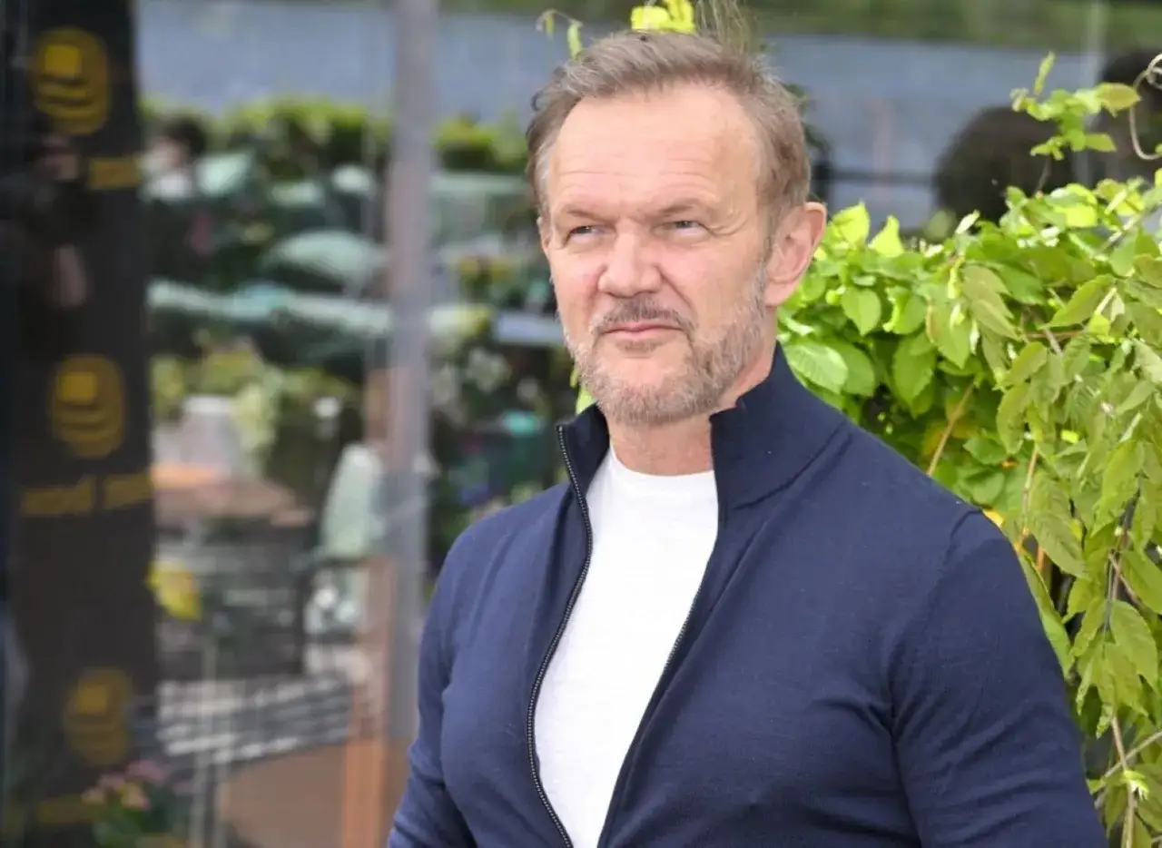 Cezary Pazura, znany aktor, w granatowym swetrze. W tle zielone liście i odbicia w szybie.