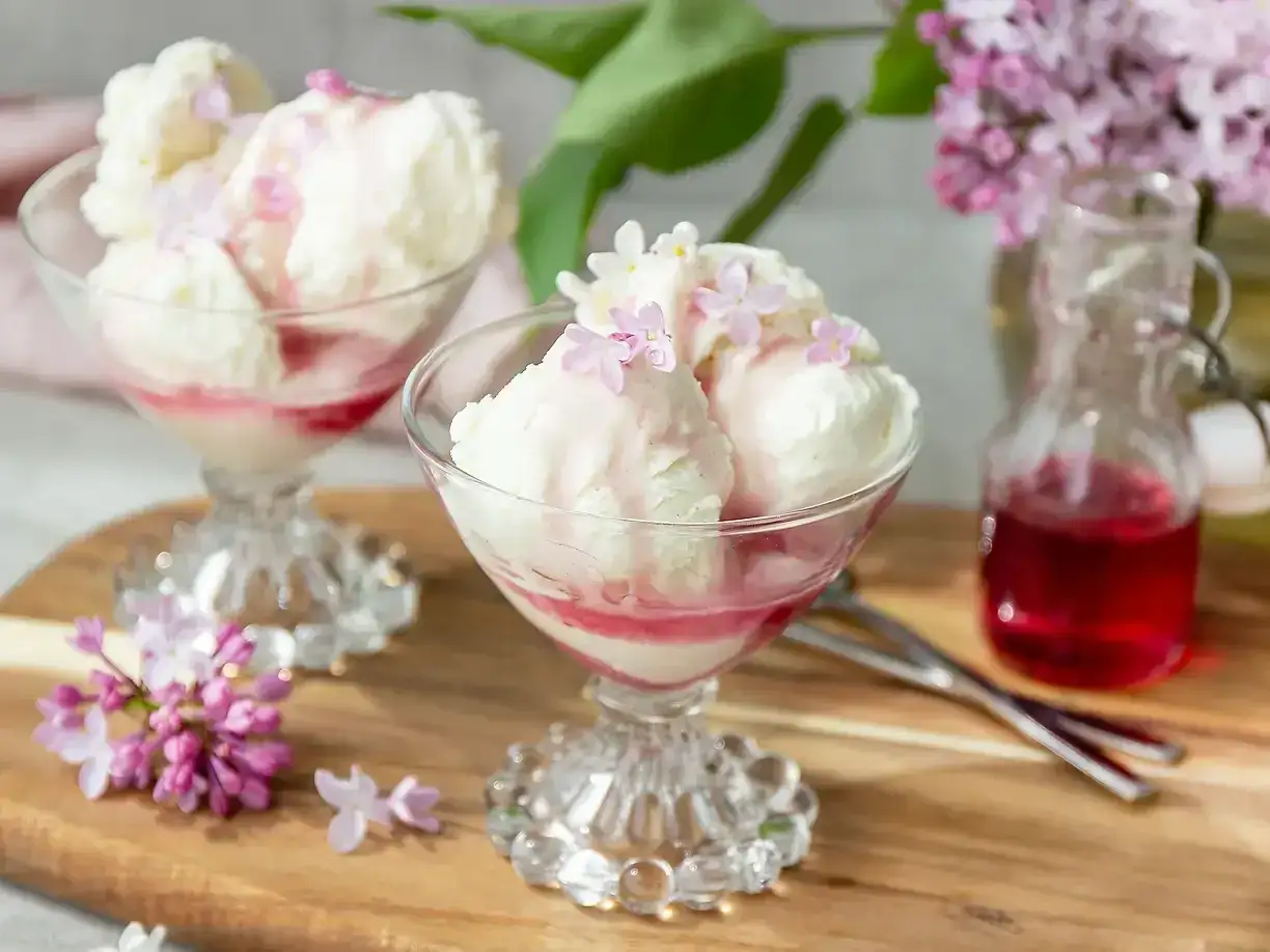 Glassb&auml;gare med vaniljglass, toppad med syrenblommor och r&ouml;d sirap. En flaska med sirap och syrenkvistar i bakgrunden. Ett h&auml;rligt syrensirap recept!