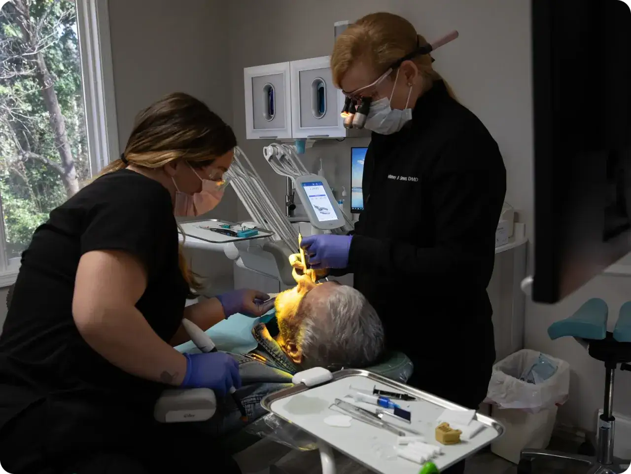 Dentystka i asystentka plombują ząb pacjentowi. Czas zabiegu, czyli ile trwa plombowanie zęba, zależy od wielu czynników.