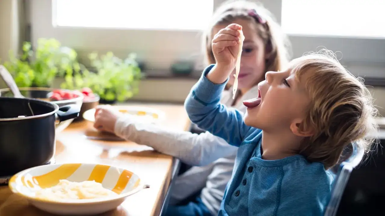 Ein Junge mit blonden Haaren isst begeistert Spaghetti, während seine Schwester ihm hilft. Sie sind so hungrig, dass die kinder essen schnell.
