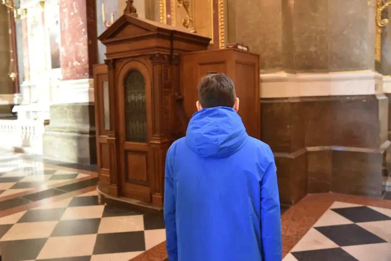 Chłopiec w niebieskiej kurtce czeka na spowiedź wielkanocną przy drewnianej konfesjonale w kościele.