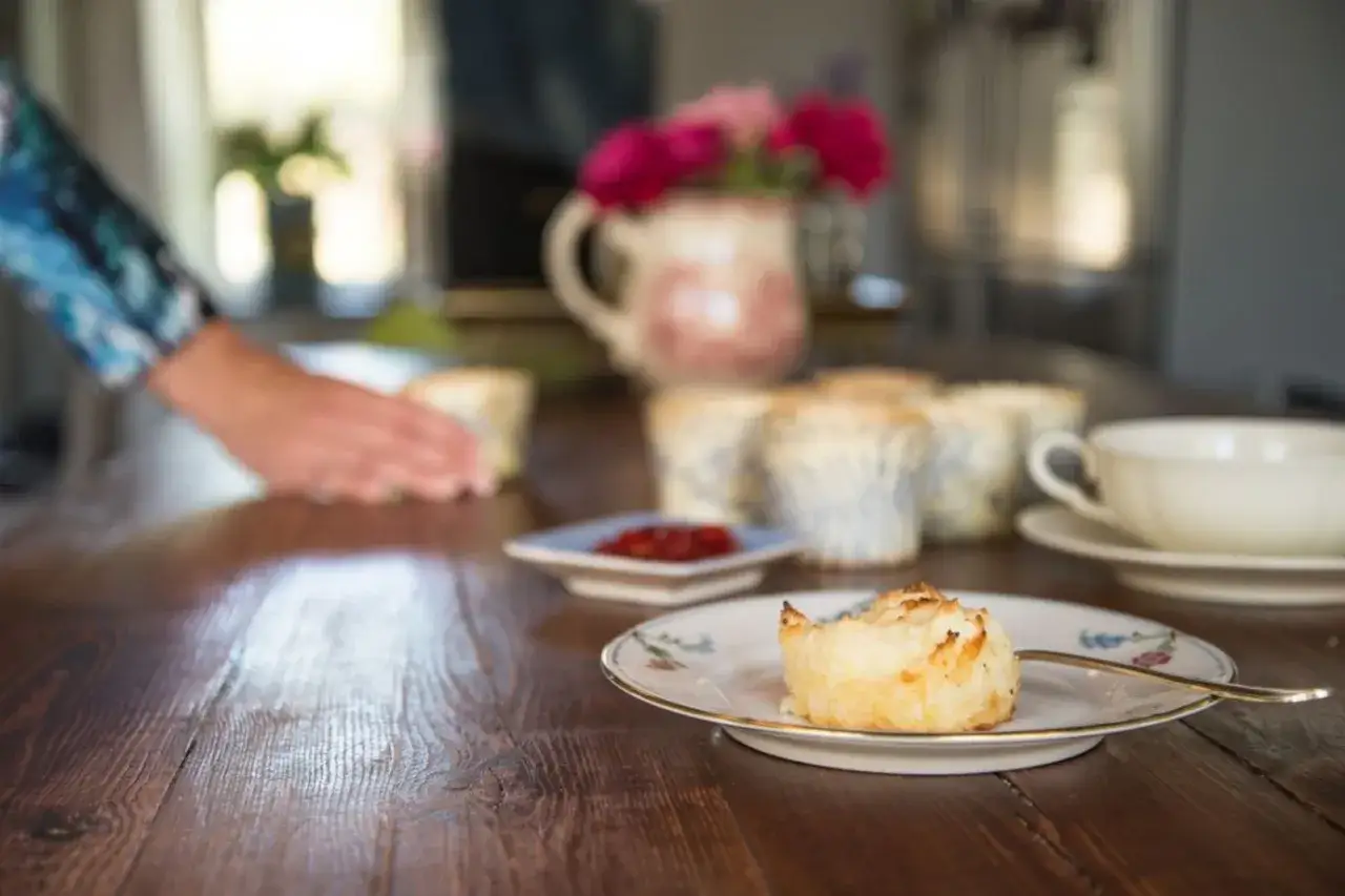 En hand lägger fram glutenfria scones på ett dukat bord. En kopp te och blommor i en vas skapar en mysig stämning.