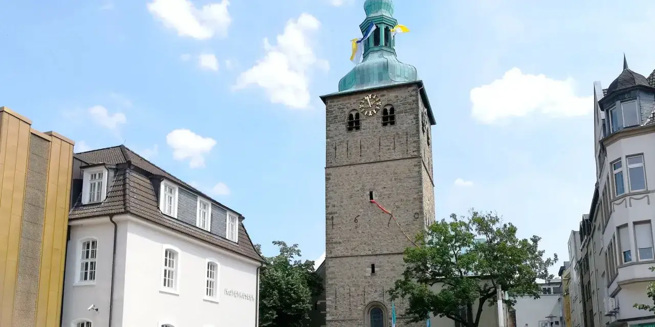 Der **Kirchplatz Recklinghausen** zeigt einen steinernen Kirchturm mit Uhr und Fahnen unter blauem Himmel. Daneben ein wei&szlig;es Geb&auml;ude.
