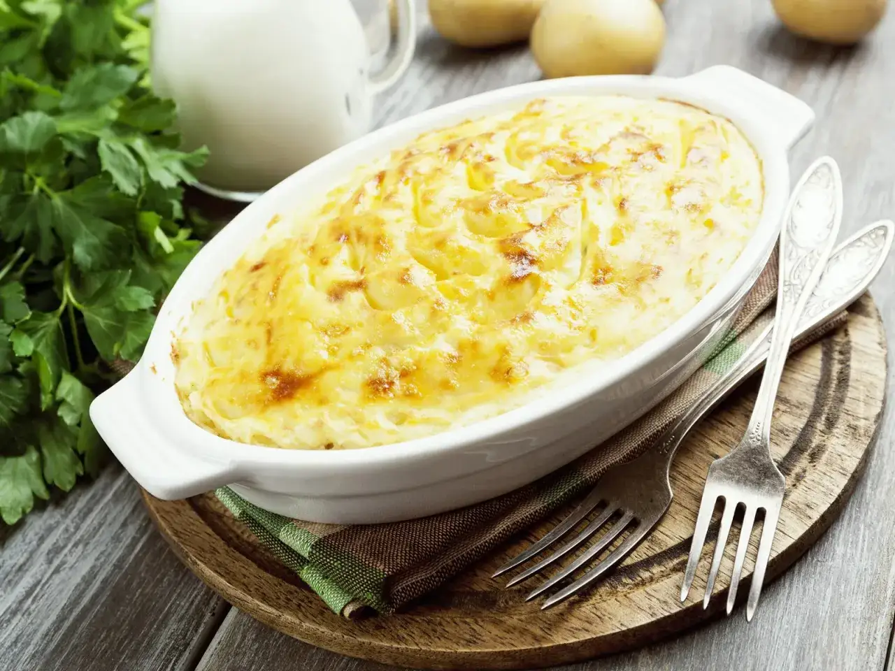 Goldbraunes, überbackenes Kartoffelgratin ohne Sahne in einer weißen Auflaufform, daneben Petersilie und Gabeln.