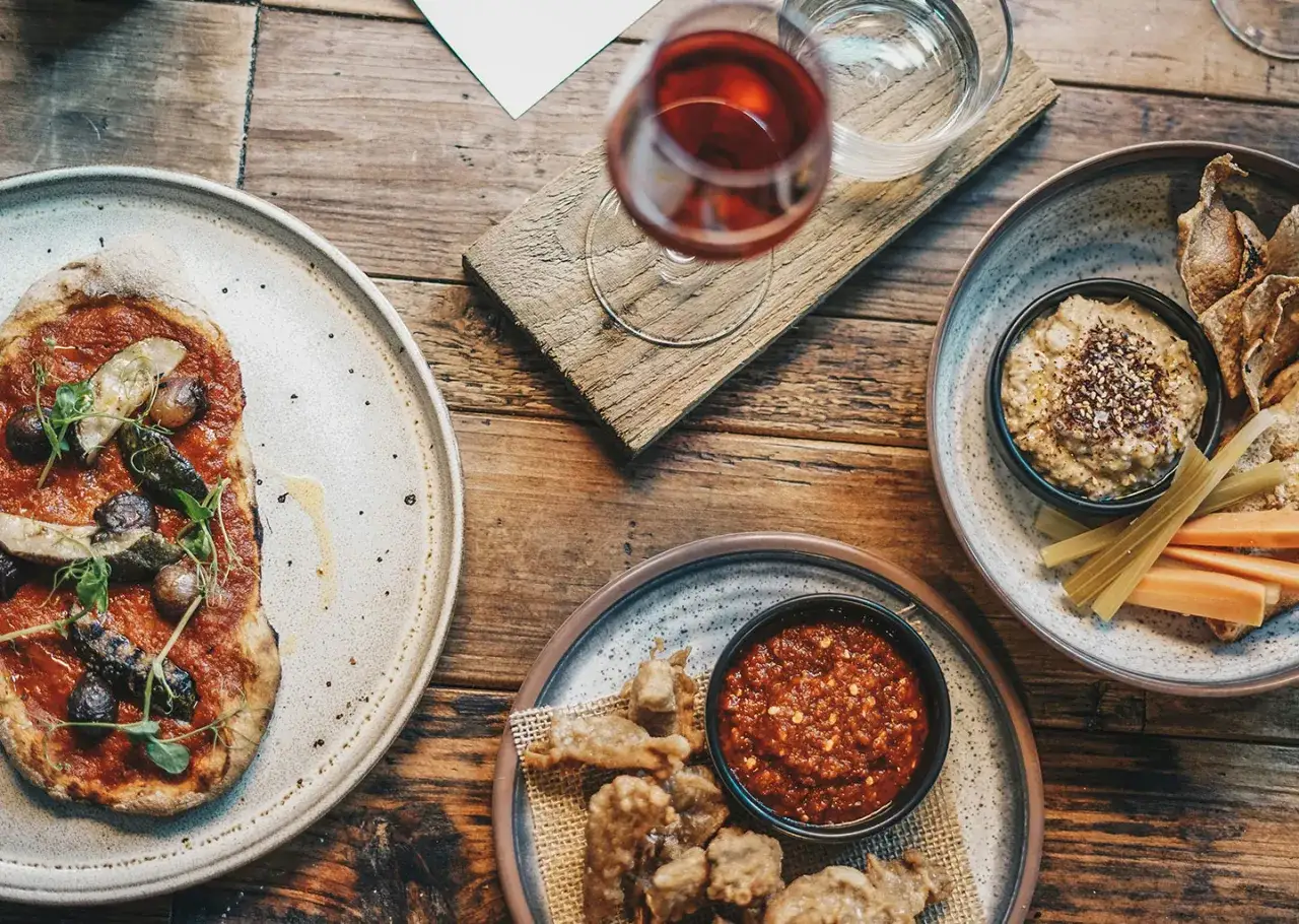Un repas qui allie savoir et mets : une pizza aux légumes, houmous et crudités, et des beignets avec sauce piquante. Un verre de vin rouge complète cette scène gourmande.