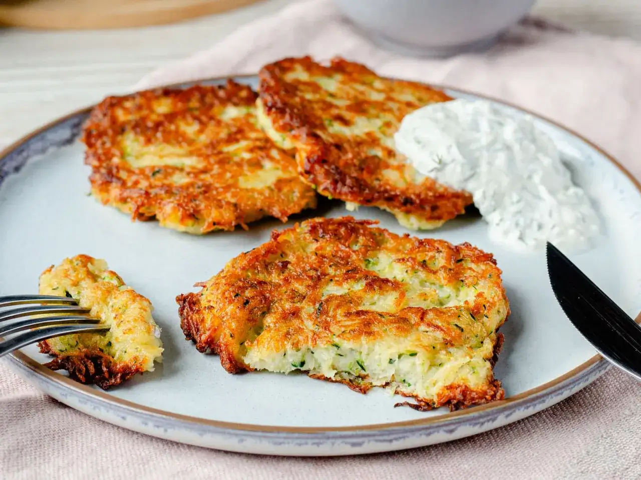 Goldbraune zucchinipuffer mit kartoffeln, knusprig gebraten und mit einem Klecks Dip serviert. Ein Gabelstück zeigt die saftige Mitte.