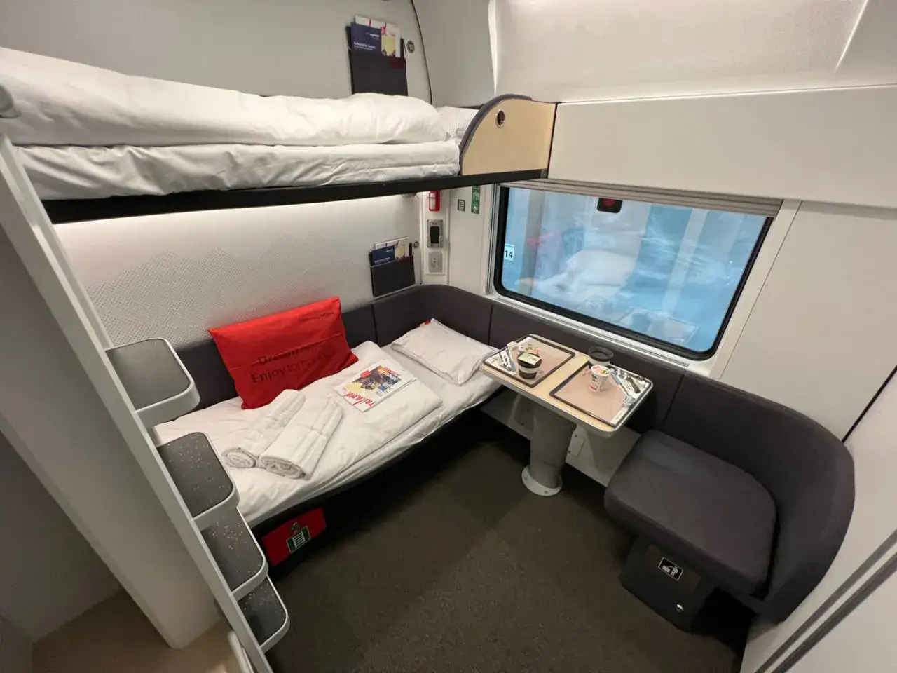 En sovhytt p&aring; ett Nightjet-t&aring;g med v&aring;ningss&auml;ngar, en bekv&auml;m sittgrupp och ett bord med frukost.