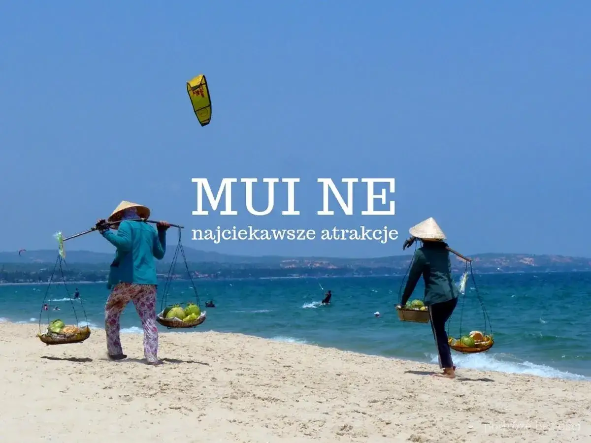Dwie kobiety w stożkowych kapeluszach niosą kosze z owocami na plaży Mui Ne. Na niebie lata żółty latawiec.
