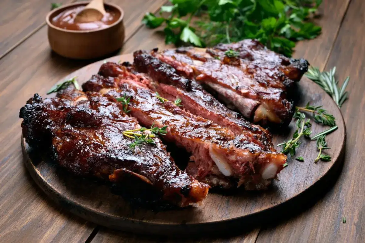 Soczyste żeberka z grilla, błyszczące od sosu, ozdobione ziołami. Obok miseczka z sosem BBQ.