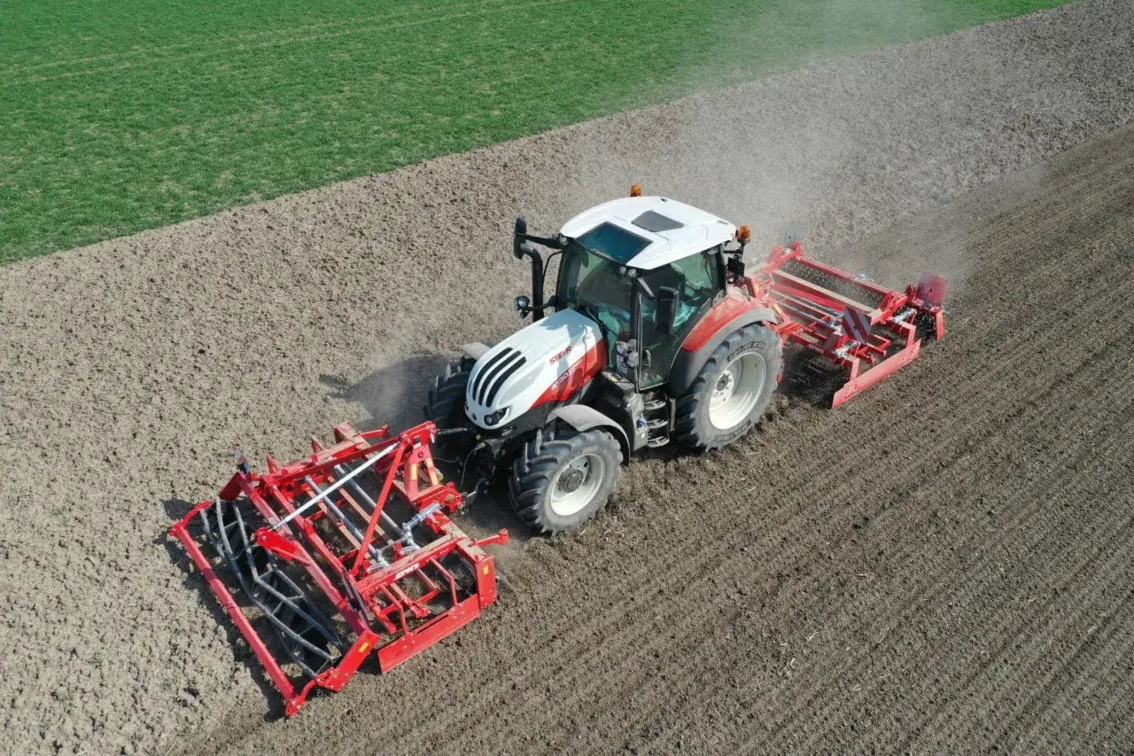 Tracteur rouge et blanc travaillant un champ labouré, préparant le sol pour un semis de prairie.