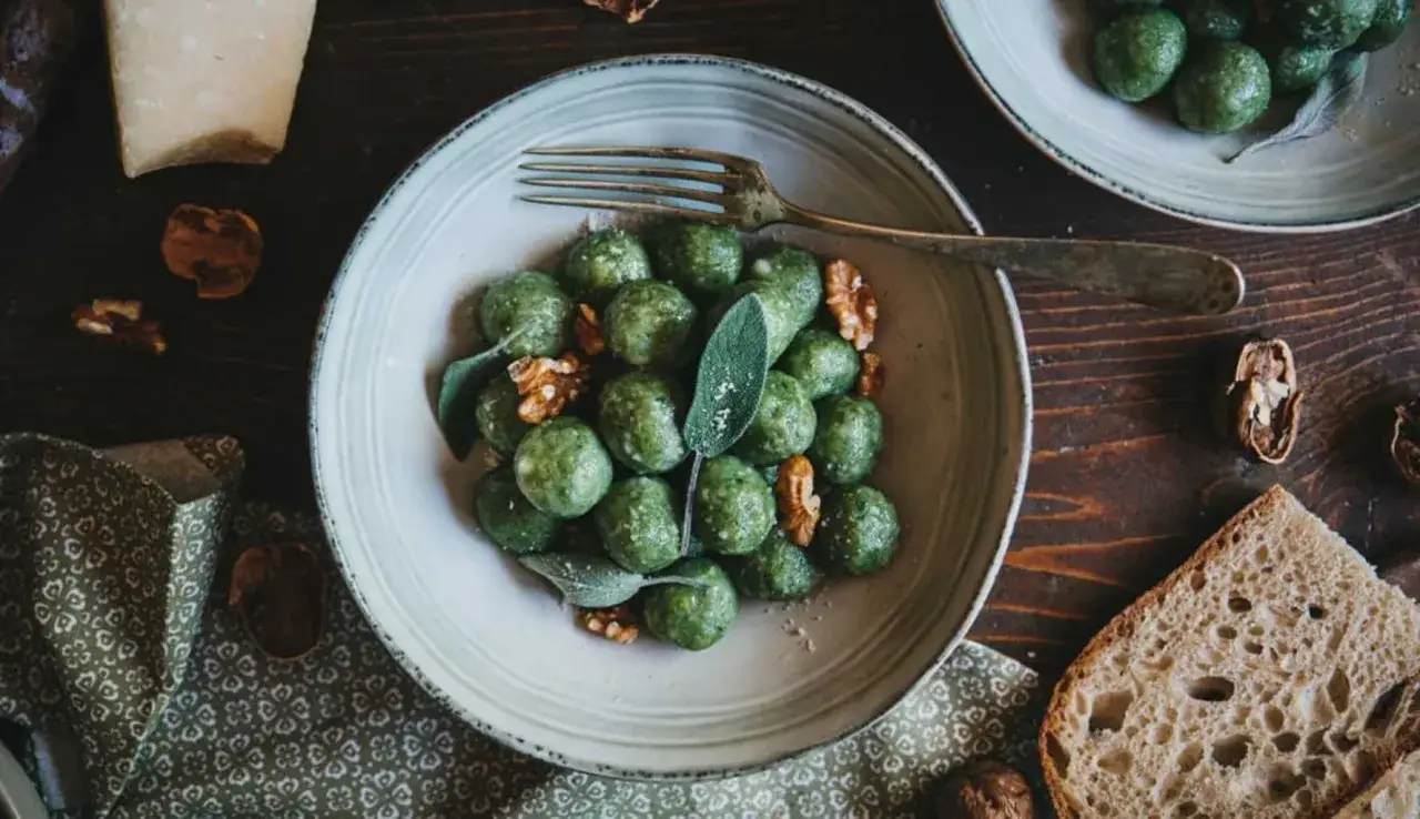 Gnocchi di pane verdi, guarniti con noci e salvia, su un piatto rustico. Accanto, pane casereccio e un pezzo di formaggio.