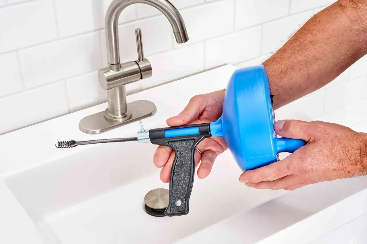 Hands hold a drain snake, ready to tackle how to clean a sink drain. The tool's coiled end is near the sink basin.