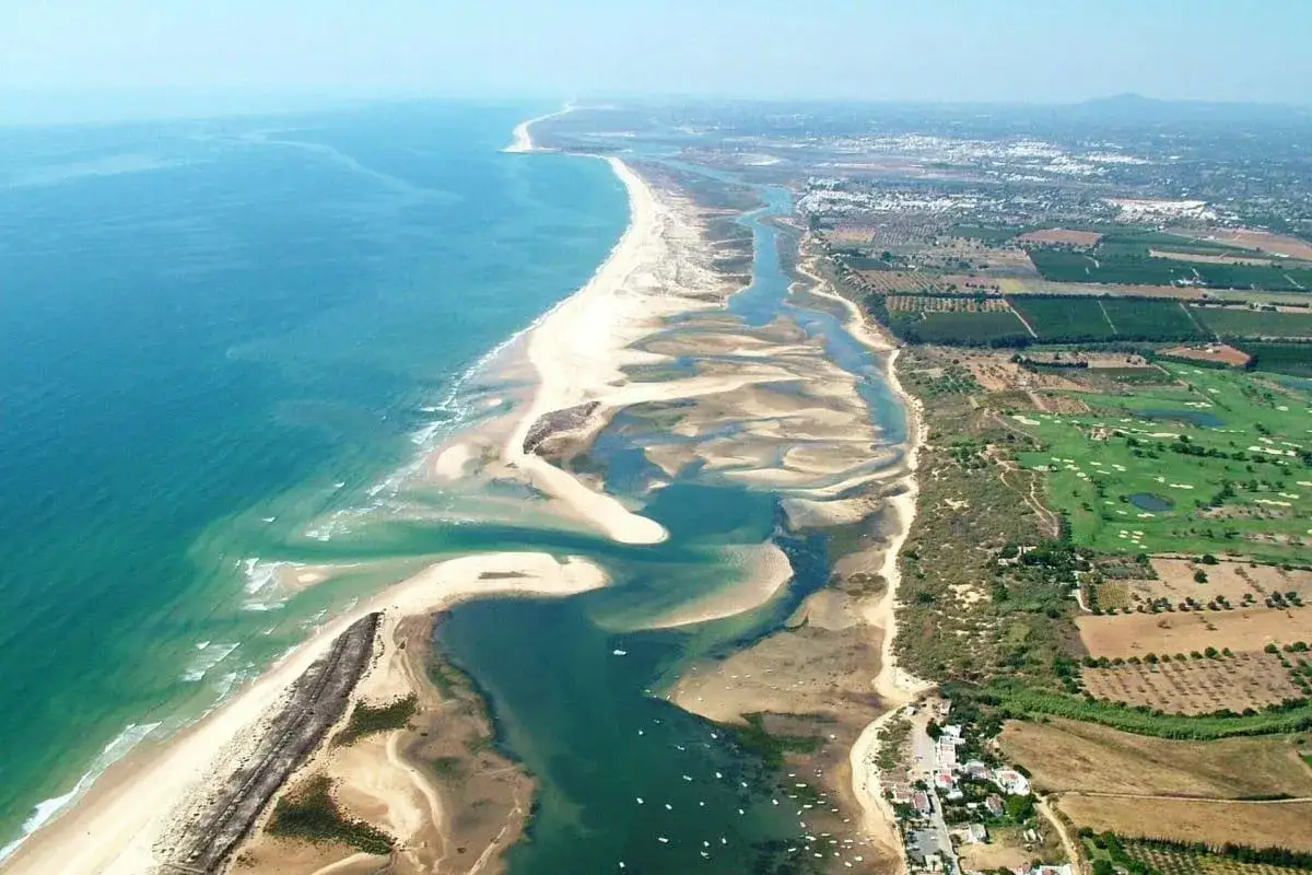 Luftaufnahme der Ria Formosa mit ihren Sandbänken, Lagunen und dem Meer. Im Hintergrund sind Felder und ein Golfplatz zu sehen.