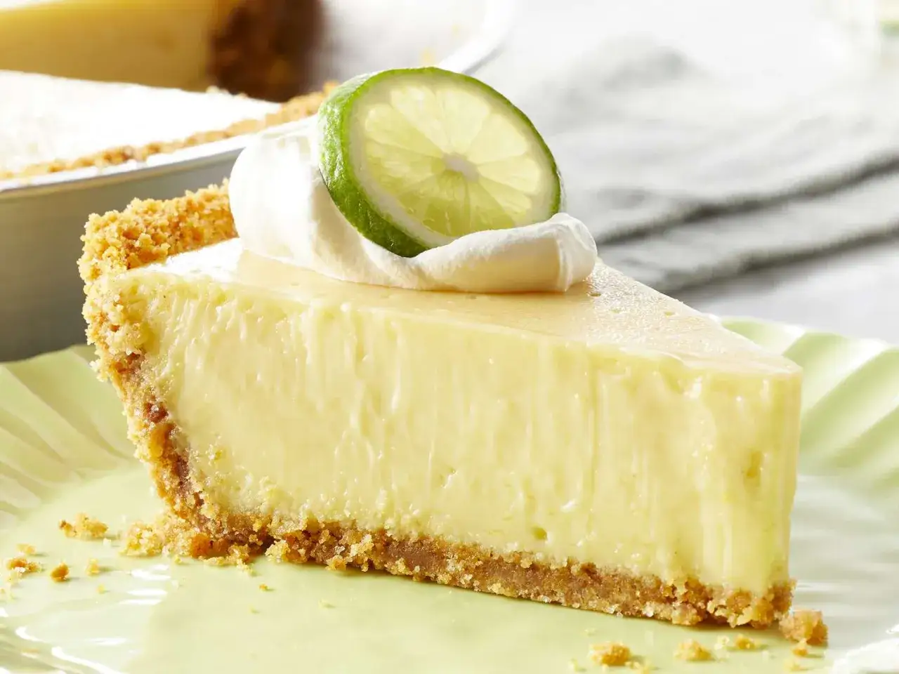Ein cremiges Stück Key Lime Pie mit Schlagsahne und Limettenscheibe auf einem hellgrünen Teller.