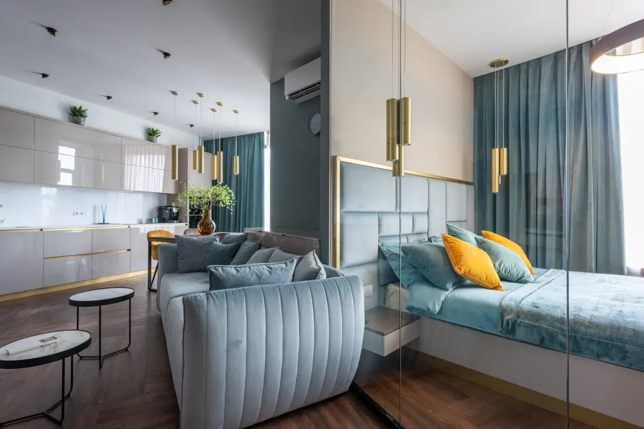 Stylowa mikrokawalerka z kuchnią, salonem i sypialnią oddzieloną szklaną ścianą. Jasne meble, błękitna sofa i łóżko.