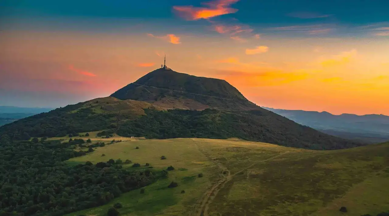 Vue du Puy de Dôme au coucher du soleil, avec un ciel orangé et des nuages. Un camping-car pourrait être garé sur les pentes verdoyantes.