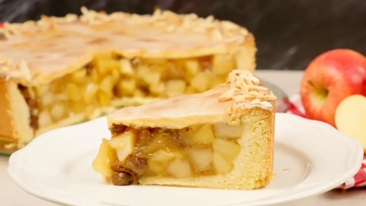 Ein Stück gedeckter Apfelkuchen mit Mandelsplittern und Zuckerguss auf einem Teller, daneben ein ganzer Kuchen und Äpfel.