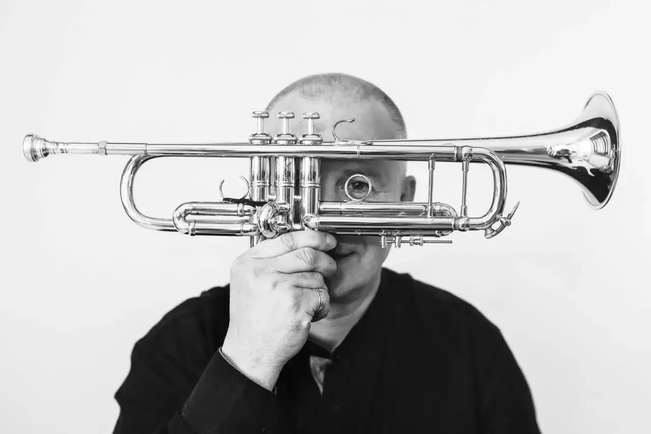 Piotr Wojtasik, muzyk, trzymający przed twarzą błyszczącą trąbkę. Jego oko patrzy przez jeden z otworów instrumentu.