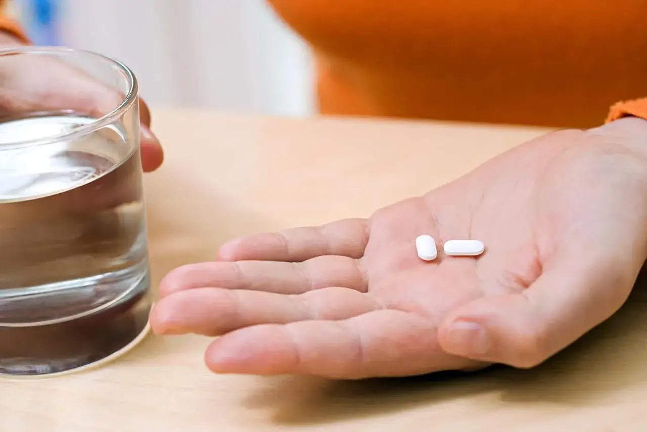 Une main tient un verre d'eau et deux comprim&eacute;s, pr&ecirc;ts pour un anti inflammatoire avant sport.