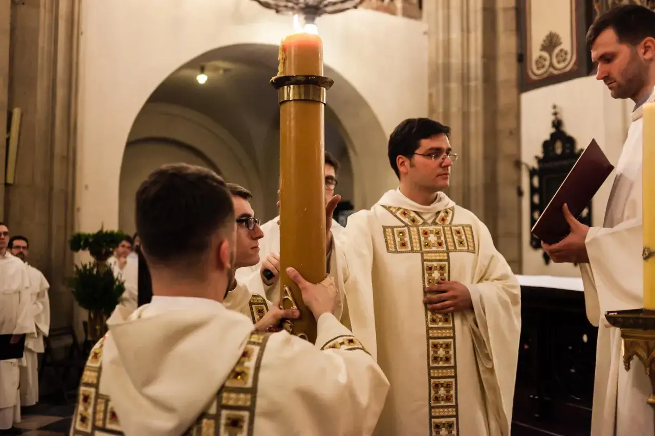 Księża w szatach liturgicznych niosą wielką świecę podczas nabożeństwa. Atmosfera sugeruje uroczystość, być może związana z czwartkiem wielkanocnym.