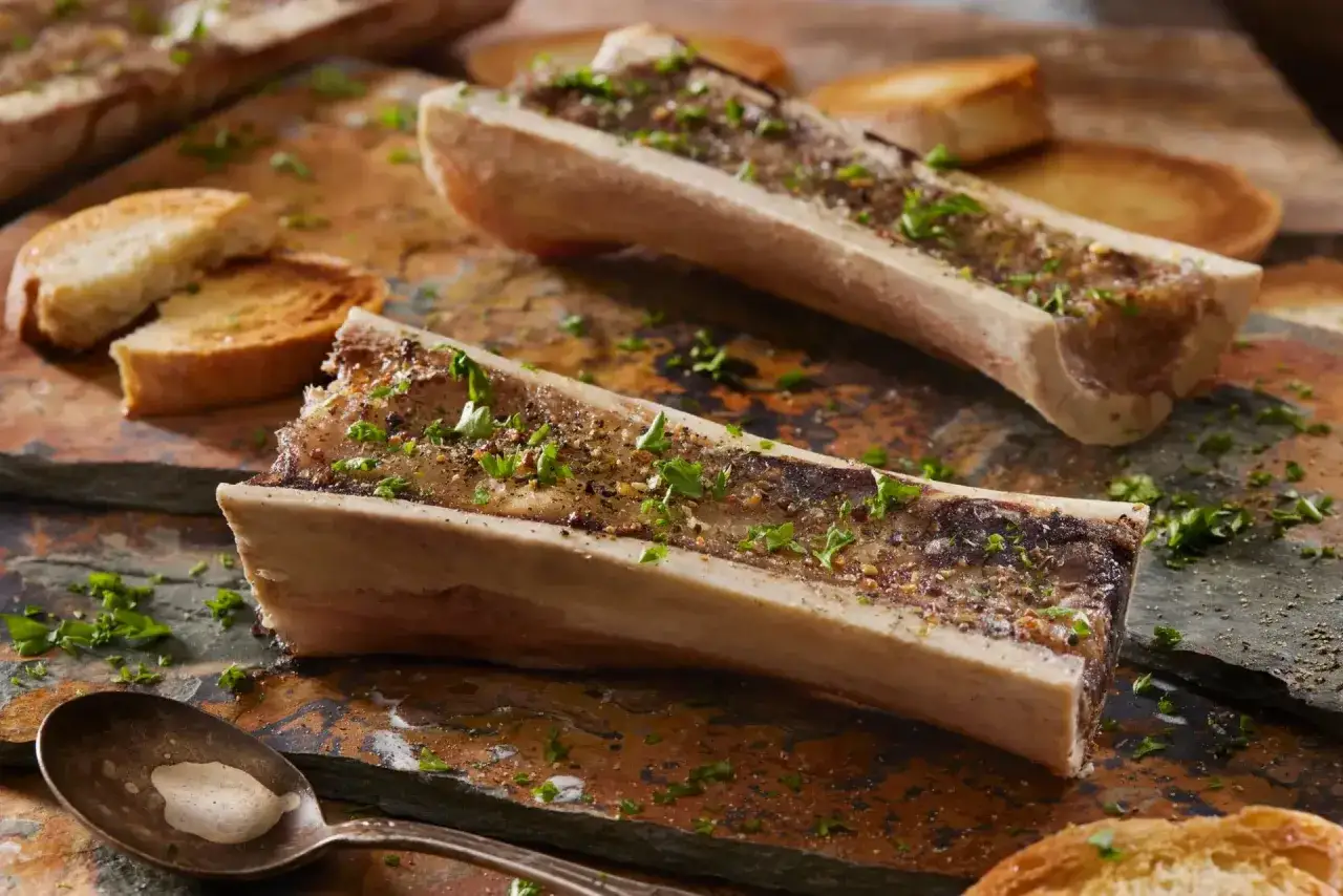 Deux os à moelle rôtis, parsemés de persil et de poivre, accompagnés de toasts grillés et d'une cuillère.