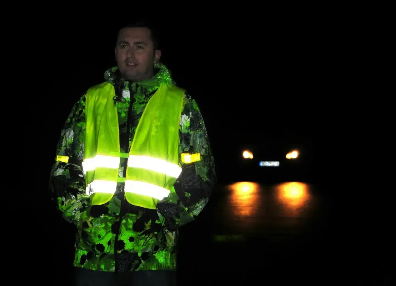 Mężczyzna w odblaskowej kamizelce i kurtce nocą. Czy kamizelka odblaskowa jest obowiązkowa? Warto ją mieć, by być widocznym dla samochodów.