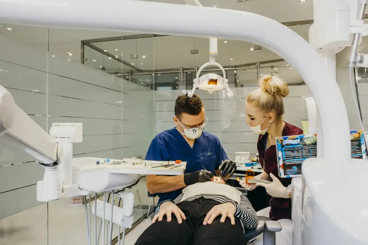 Dentysta i asystentka w masce pracują przy pacjencie w gabinecie stomatologicznym.
