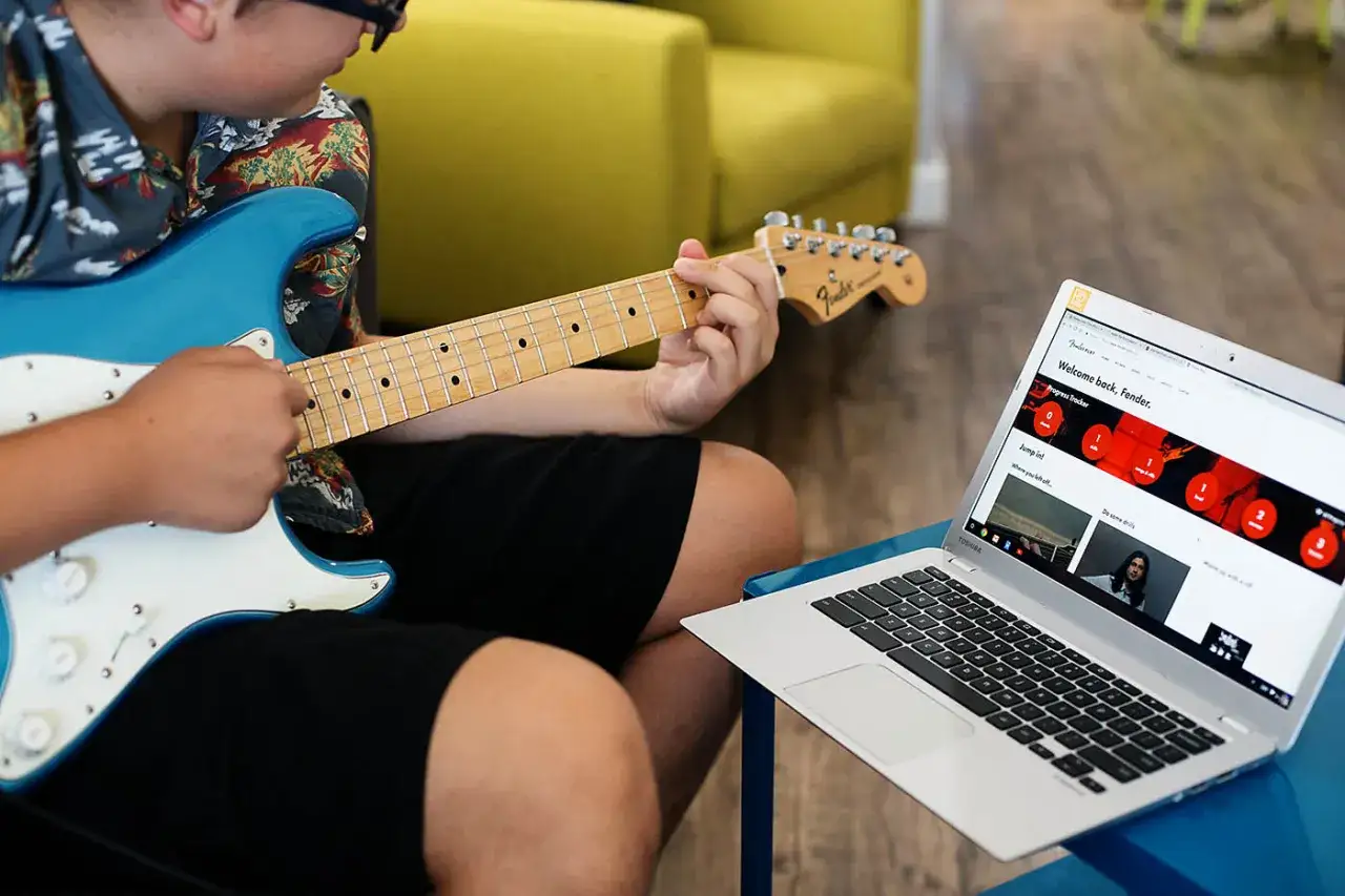 Junge lernt E-Gitarre online mit Fender Play. Ein blauer Fender Stratocaster liegt auf seinem Schoß, daneben ein Laptop mit der Lernplattform.